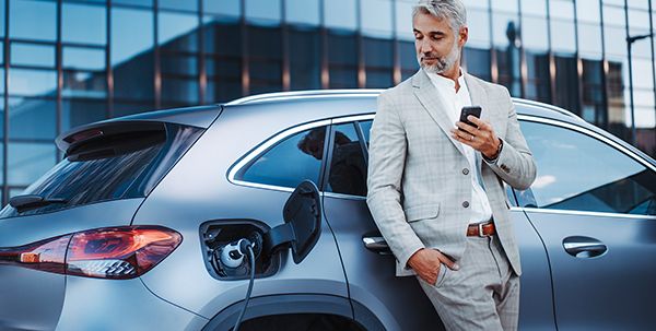 Image of businessman holding smartphone while charging car at electric vehicle charging station, closeup.
